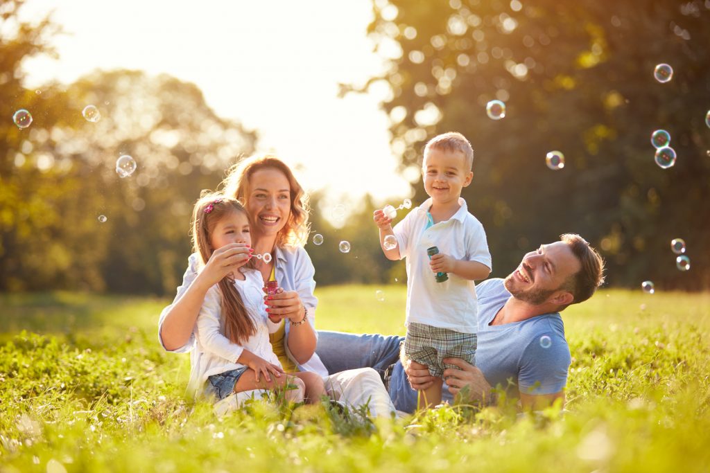 A family outside in the grass, the children are blowing soap bubbles and the family is laughing and having a great time together.